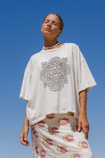 Shanti Tee White — front view upper body against blue sky, white oversized cotton tee with charcoal hand-printed Daughters of India mandala logo, paired with peony floral skirt | Daughters of India