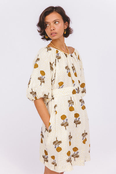 Dahlia Mini Dress Marigold — studio model side glance with hand in pocket, on-shoulder tie neckline, white linen with golden marigold flower hand block print, Jaipur artisan made mini dress | Daughters of India