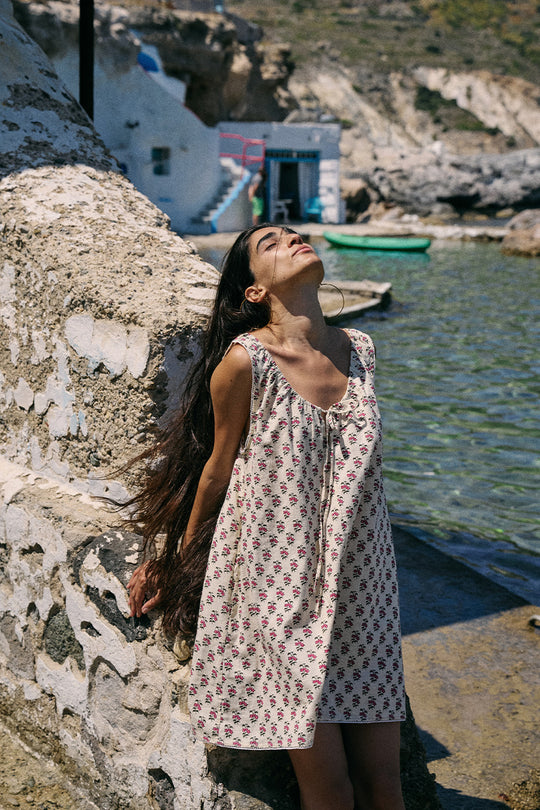 Sundress Mini Peony — model leaning against stone wall by coastal harbor looking up, hand block print peony floral cotton mini dress, Mediterranean seaside lifestyle | Daughters of India